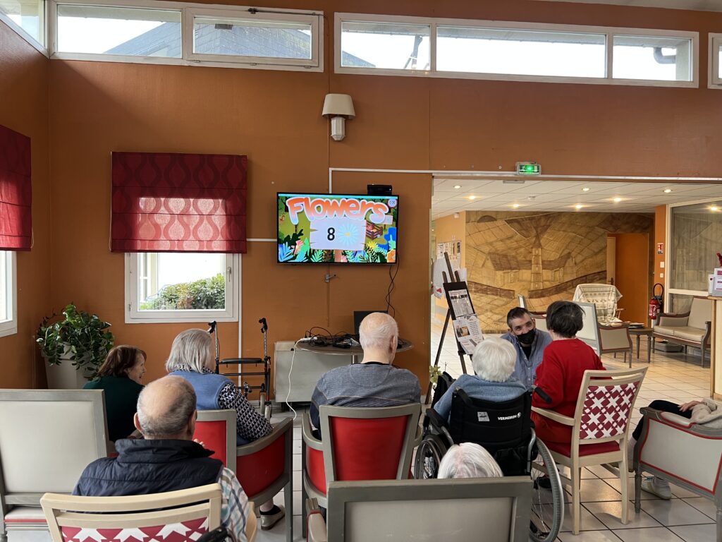 Séance d'animation collective en EHPAD où un groupe de personnes âgées, dont une en fauteuil roulant, est assis face à un écran affichant un jeu interactif 'Flowers'. Un animateur en tenue bleue se tient près des participants dans une salle spacieuse aux murs orange terracotta avec grandes fenêtres hautes. L'espace moderne dispose de fauteuils confortables rouge et blanc, et s'ouvre sur d'autres zones communes lumineuses en arrière-plan.