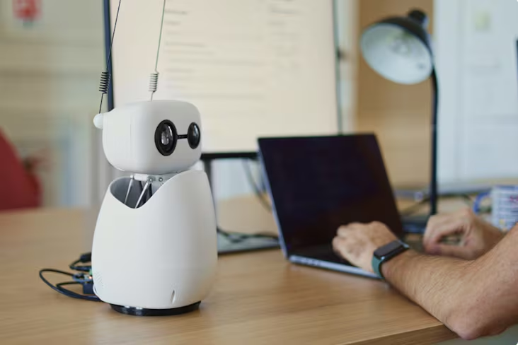 Reachy Mini, un robot humanoïde de poche, contrôlé par intelligence artificielle, développé par Pollen Robotics, blanc avec des yeux noirs, posé sur un bureau à côté de quelqu'un à un ordinateur